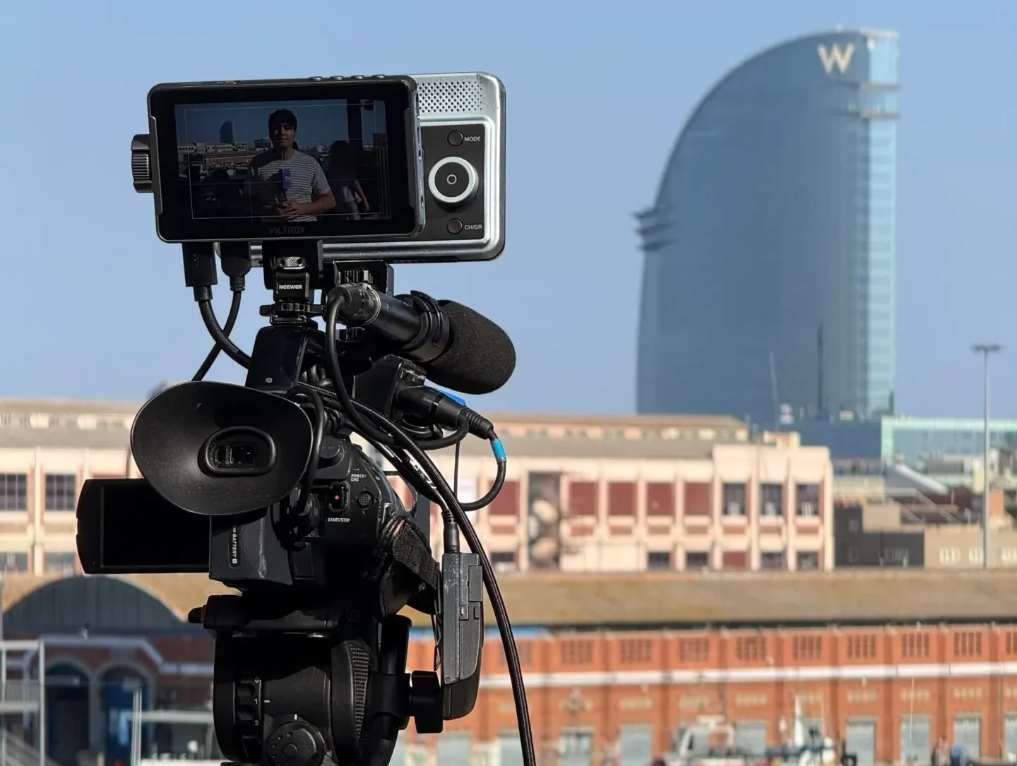 Cámara de vídeo profesional en trípode grabando con el Hotel W y el puerto de Barcelona al fondo